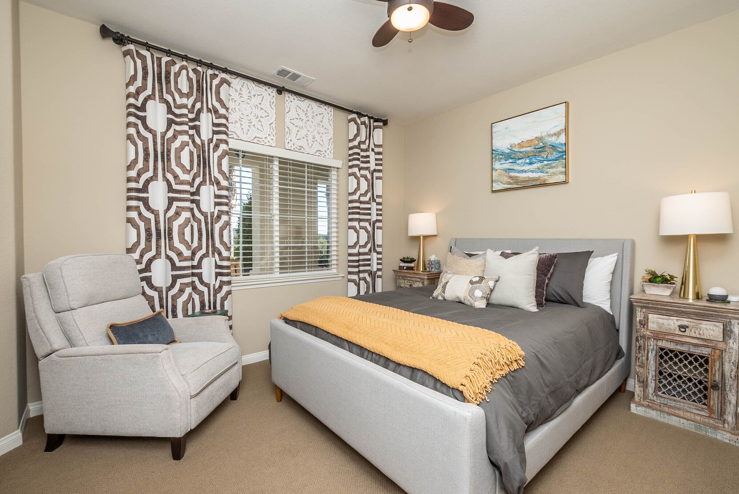 A bedroom with a bed, a chair, a nightstand, a lamp, and a ceiling fan.