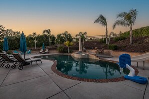 A swimming pool with lounge chairs and umbrellas, surrounded by palm trees and a fence. - Dream Getaway:Putting Green, Game Room, Volleyball (Valley Center)