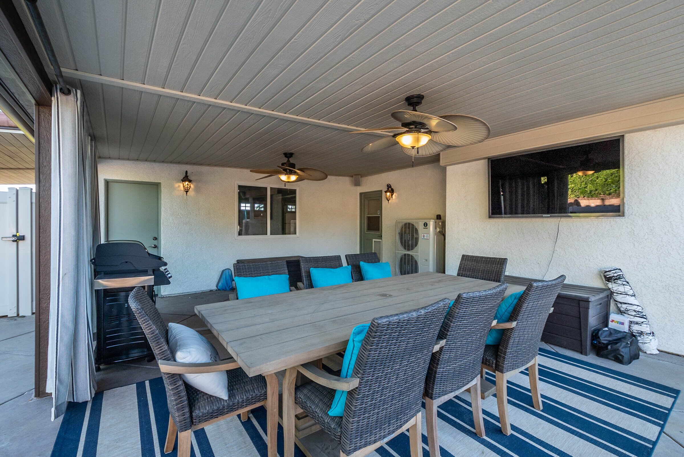 Une terrasse extérieure avec une table en bois et des chaises en osier, des ventilateurs de plafond et un barbecue.