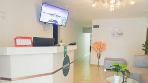 Zona de recepción de un hotel con un televisor de pantalla plana en la pared, un mostrador de recepción con un calendario, una planta en maceta y una zona de estar con un sofá y una mesita.