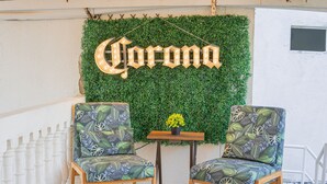 Dos sillas con un estampado de hojas, una mesita con una planta en maceta y una pared con un letrero de 'Corona'.