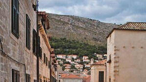 Una calle estrecha con mesas y sillas en la terraza, edificios de piedra y montañas al fondo.