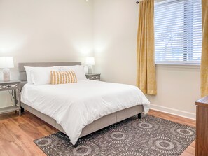 A neatly made bed with white bedding and a patterned pillow, flanked by bedside tables with lamps, a patterned rug, and a window with curtains.