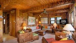 L’intérieur d’un chalet en bois est aménagé avec un ensemble de canapés, deux lampes, un ventilateur de plafond et une enseigne sur laquelle on peut lire : « Vacances et ventes à Coastal Carolina ».
