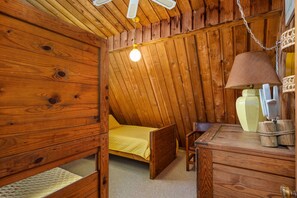 Une pièce en bois avec un plafond en pente, un lit recouvert d’une couverture jaune, une table avec une lampe et une chaise.