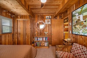 A wooden cabin interior with a bed, a bookshelf, a mirror, and a framed picture on the wall.