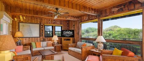 A cozy wooden cabin interior with a sofa, chairs, a television, and a large window offering a view of the forest.