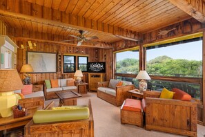 A cozy wooden cabin interior with a sofa, chairs, a television, and a large window offering a view of the forest. - Dunesbury:Oceanfront, dog-friendly, four bedrooms, mini-vacation anytime! (Corolla)
