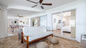 A modern living room with a white sofa, two round ottomans, a ceiling fan, and a large sliding glass door leading to a bedroom. - Picturesque Watefront, Heated Pool, Ping Pong Table - Villa Sunset Riverhouse - Roelens Vacations (Cape Coral)