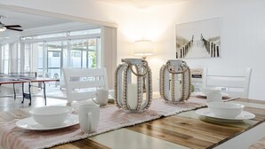 A dining area with a wooden table, white chairs, and a white table runner. There are two white lanterns with bead trim and a wall-mounted lamp. A framed picture hangs on the wall. - Picturesque Watefront, Heated Pool, Ping Pong Table - Villa Sunset Riverhouse - Roelens Vacations (Cape Coral)