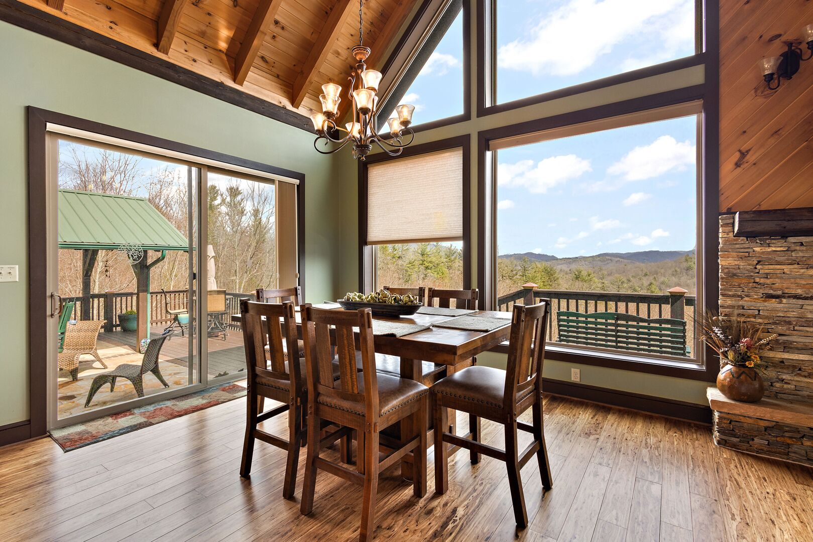 A dining area with a wooden table and chairs, large windows with mountain views, and a wooden ceiling.