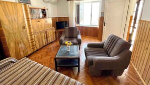 Een gezellige woonkamer met een bank, fauteuil en een salontafel met een vaas. Er staat een televisie op een houten kast en een raam met gordijnen.