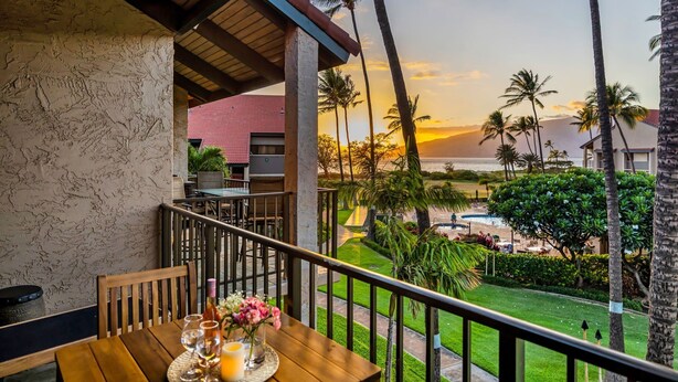 Ein Balkon mit Holztisch, Stühlen und einer Vase mit Blumen. Im Hintergrund ist der Blick auf das Meer und Palmen zu sehen.