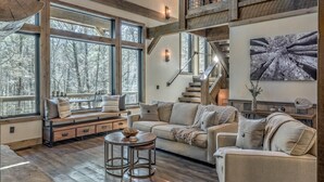 A spacious living room with a large window overlooking a snowy forest, a cozy sofa set, a small round table, and a staircase leading to an upper level. - The Otter Side - Pet Friendly | CreekFront | Mountain Modern Luxury (Epworth)