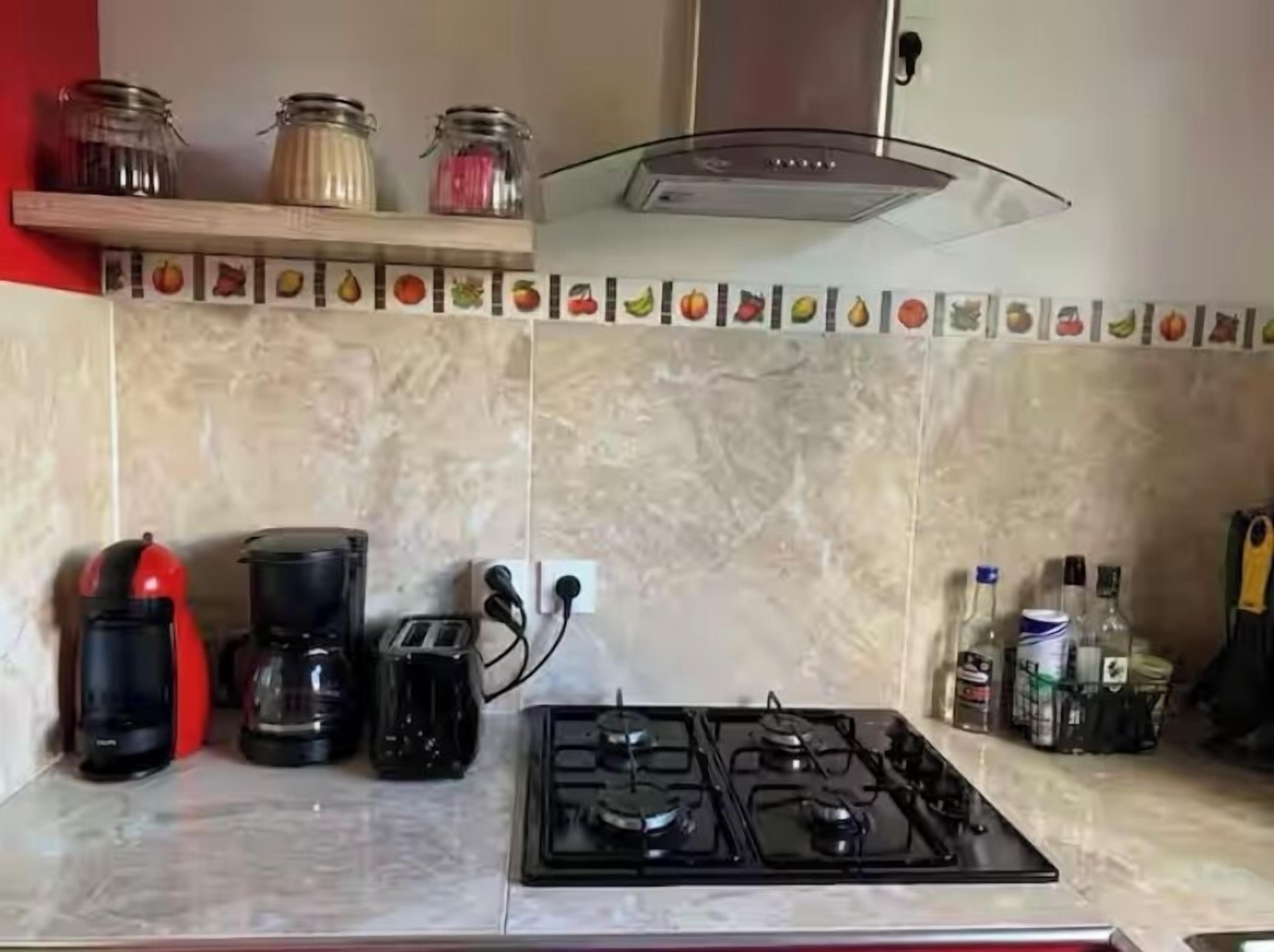 A kitchen with a gas stove, coffee maker, and a red kettle. There are jars on a shelf above the stove.