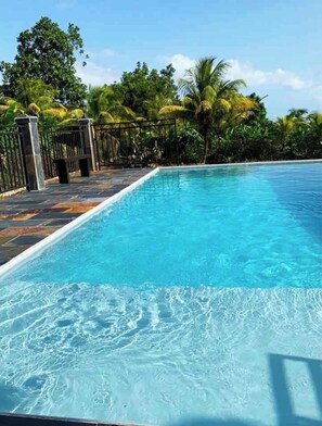 Une piscine d’un bleu éclatant, bordée d’une terrasse carrelée, entourée d’une végétation luxuriante et protégée par une clôture en métal.
