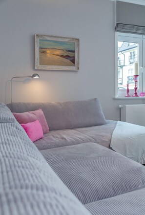 Een slaapkamer met een grijze bank, een schilderij aan de muur, een bedlampje en een raam met uitzicht op gebouwen.