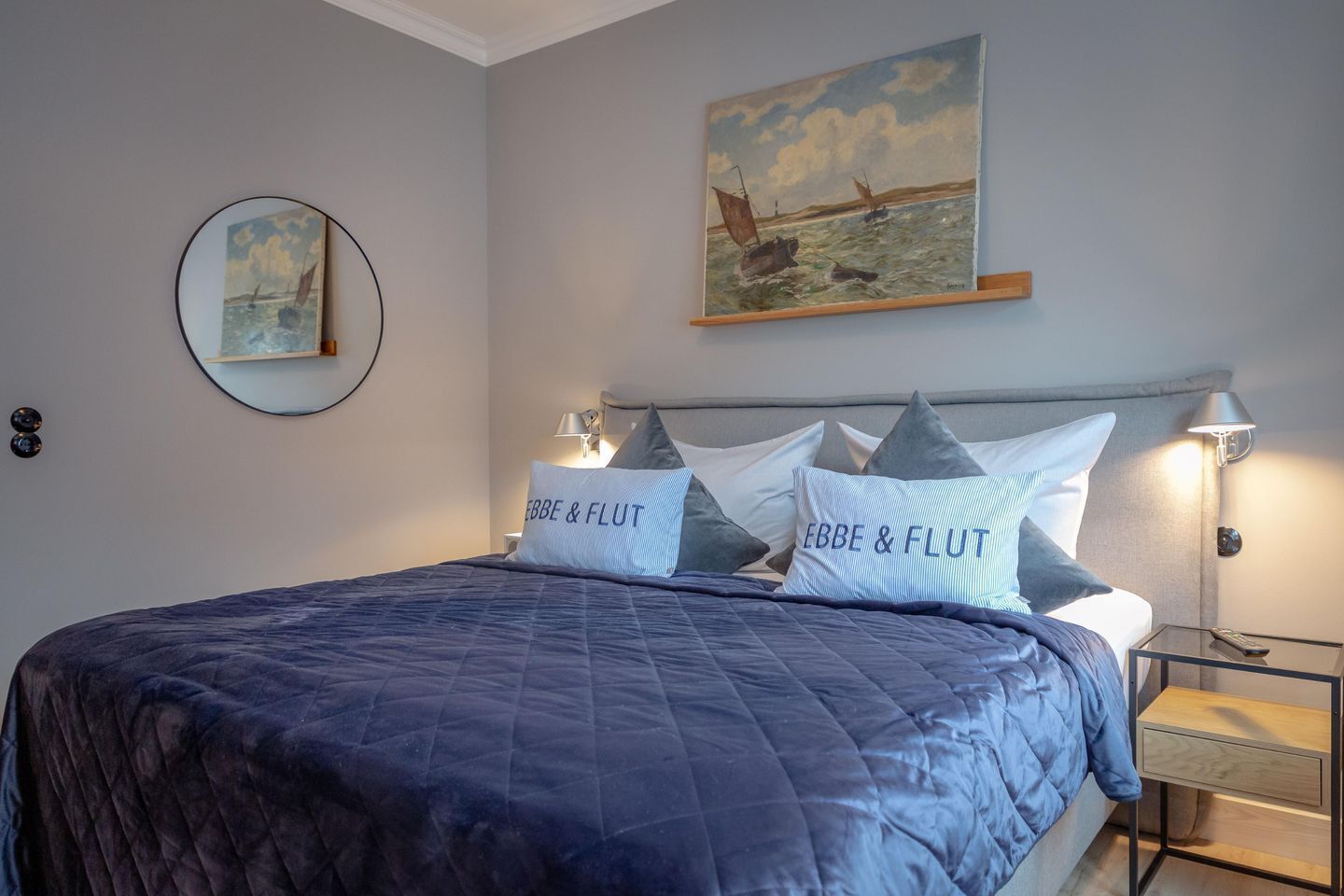 A neatly made bed with a dark blue quilt, two pillows with "EBBE & FLUT" text, a painting of boats on the wall, and a round mirror on the left.