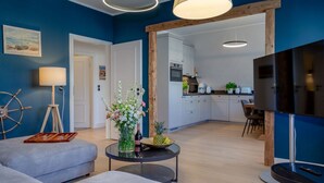 Una sala de estar moderna con un sofá, una mesa de centro y un televisor. Se observa una cocina al fondo con una zona de comedor.