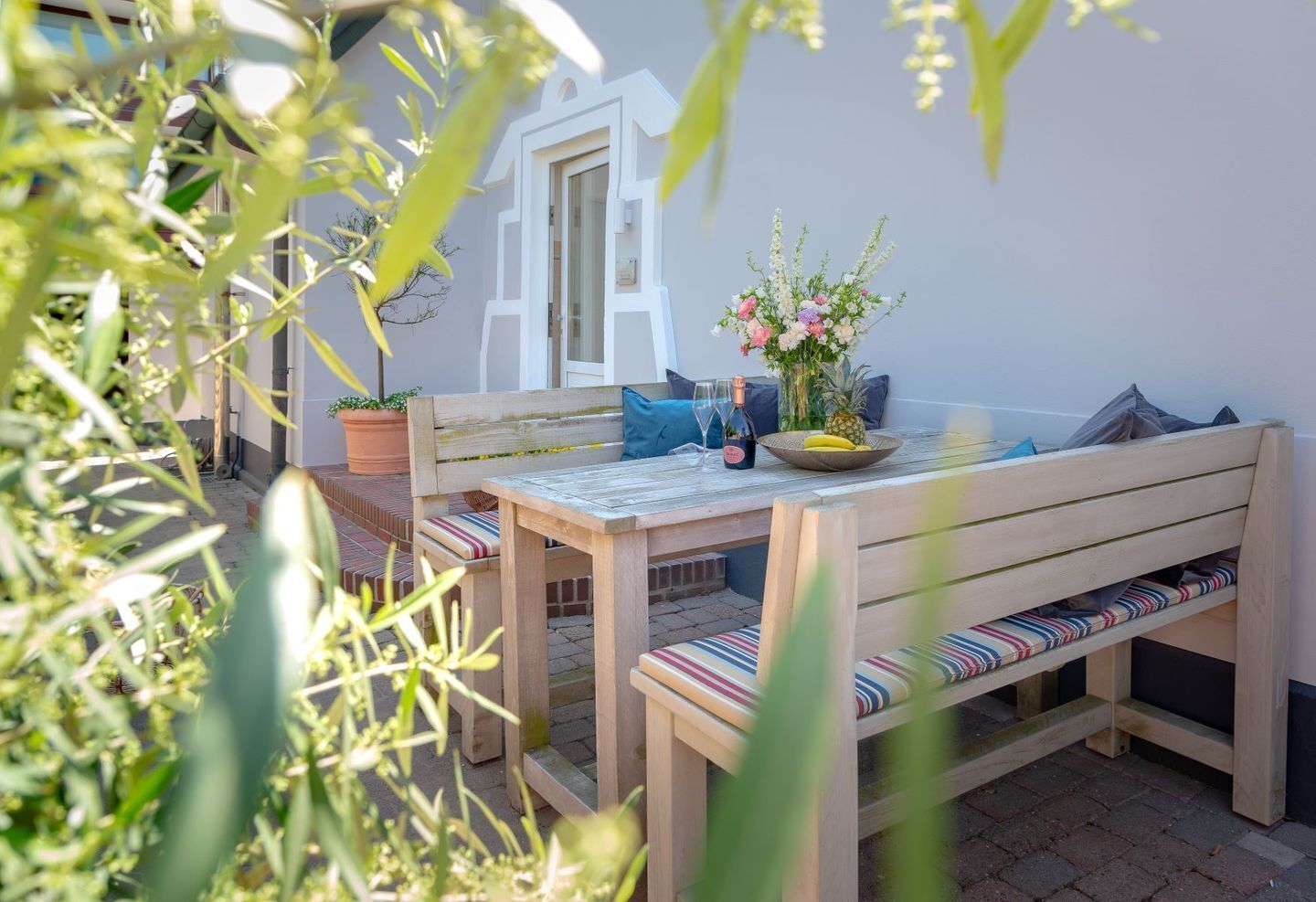An outdoor patio with a wooden table and benches, surrounded by potted plants and a white wall.