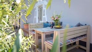 Un patio al aire libre con una mesa y bancos de madera, rodeado de plantas en macetas y una pared blanca.