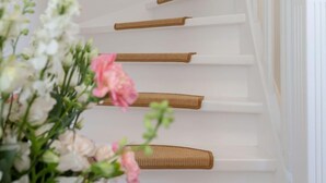 Una escalera con peldaños blancos y barandillas de madera, que conduce a un rellano con cuadros en la pared.