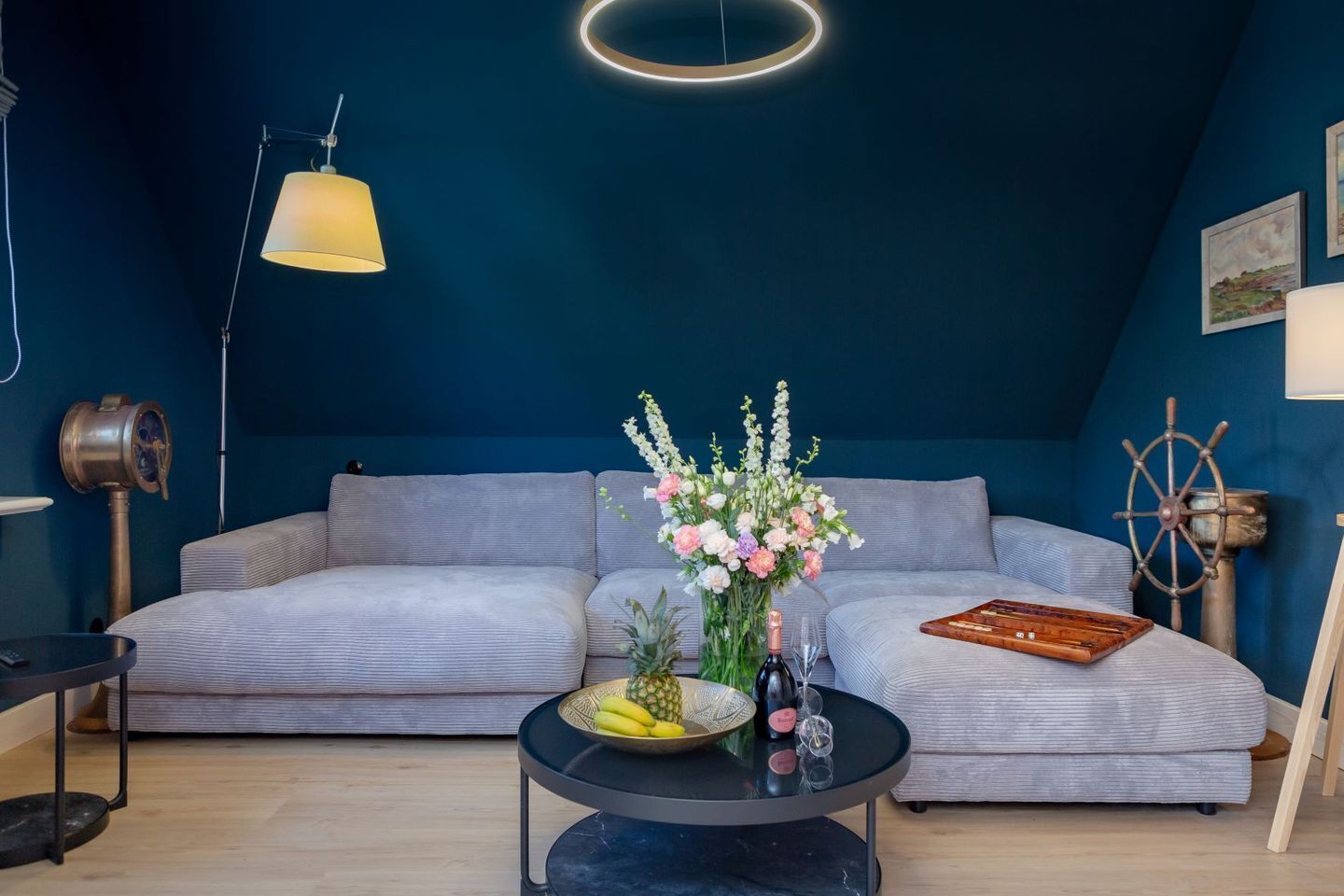 Una sala moderna con un sofá gris, una mesa de centro redonda con un centro de flores y decoración de pared temática náutica.