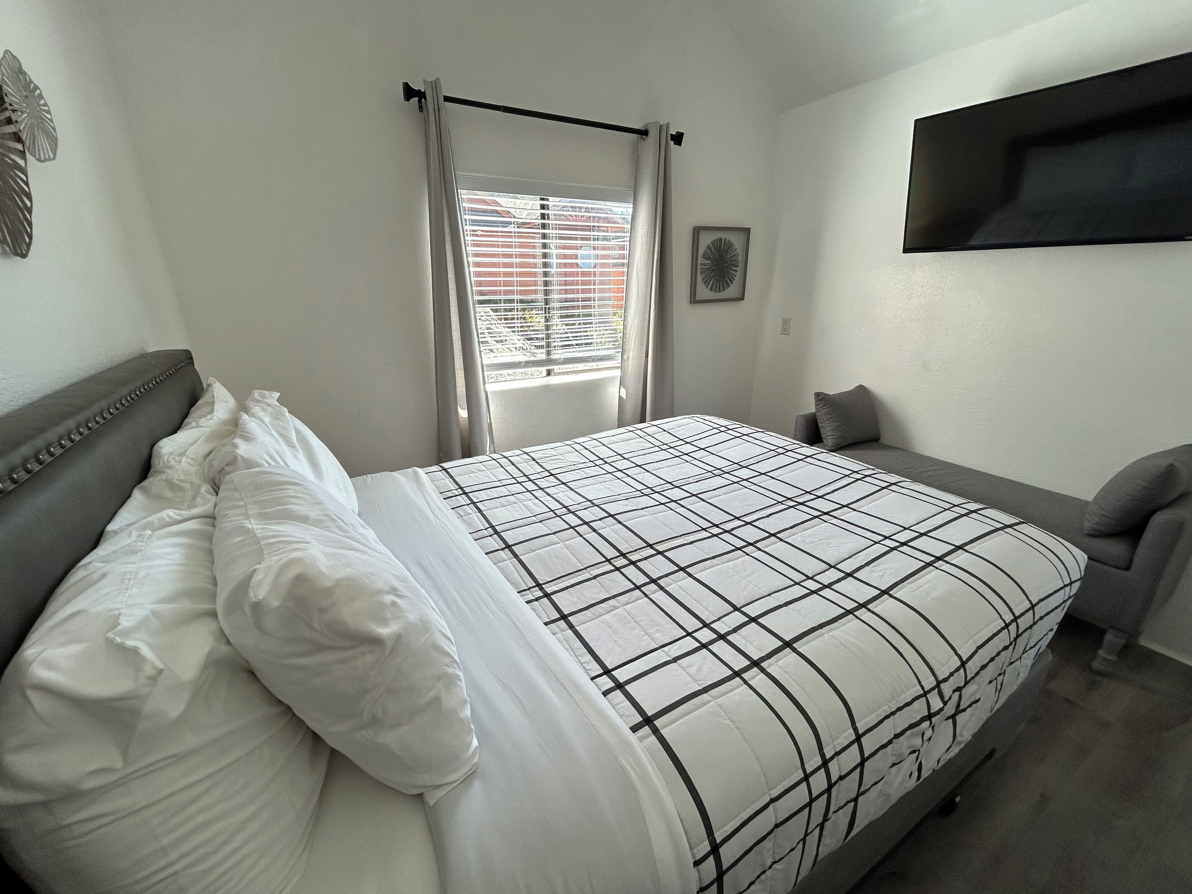 Una cama bien hecha con una colcha a cuadros, un cabecero tapizado en gris y un sillón también en gris.