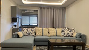 A modern living room with a grey sectional sofa, a matching ottoman, a wooden coffee table, and a fluffy rug. - Seven Heaven Plaza (Brufut)