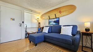 A cozy living room with a blue sofa, a surfboard wall art, a wooden coffee table, and a lamp. - 9th Floor Studio Apt, Full kitchen & Insane views! (Honolulu)