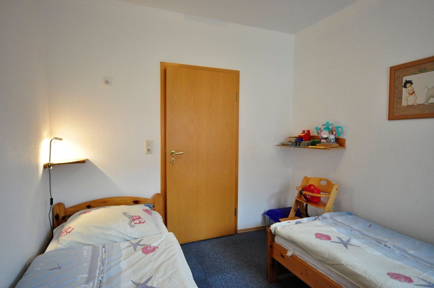 Een kleine, opgeruimde kamer met twee eenpersoonsbedden, een houten deur, een wandplank met spullen en een nachtkastje met een lamp.