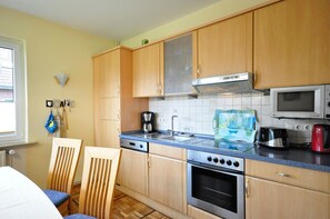 A compact kitchen with wooden cabinets, a microwave, a coffee maker, and a toaster. There is a window on the left and a chair in the foreground. - Ferienwohnung Sonnenblume - Ferienhaus Wempen (Werdum)