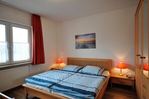A bedroom with a bed, two bedside lamps, a window with curtains, a radiator, and a painting on the wall. - Ferienwohnung Sonnenblume - Ferienhaus Wempen (Werdum)