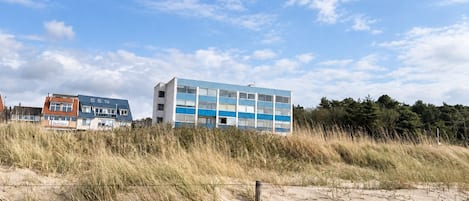 A building with a flat roof and a blue and white facade, situated on a sandy terrain with grass in the foreground.