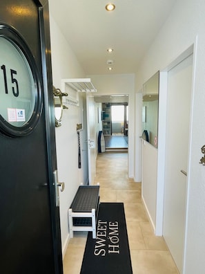 A hallway with a door labeled '15', a bench, a 'SWEET HOME' mat, and a view into a room with a bed and a TV.