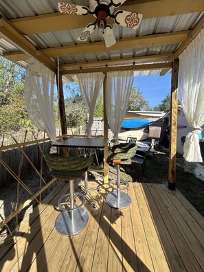 An outdoor patio with a wooden deck, two green cushioned chairs, a metal table, and a ceiling fan.