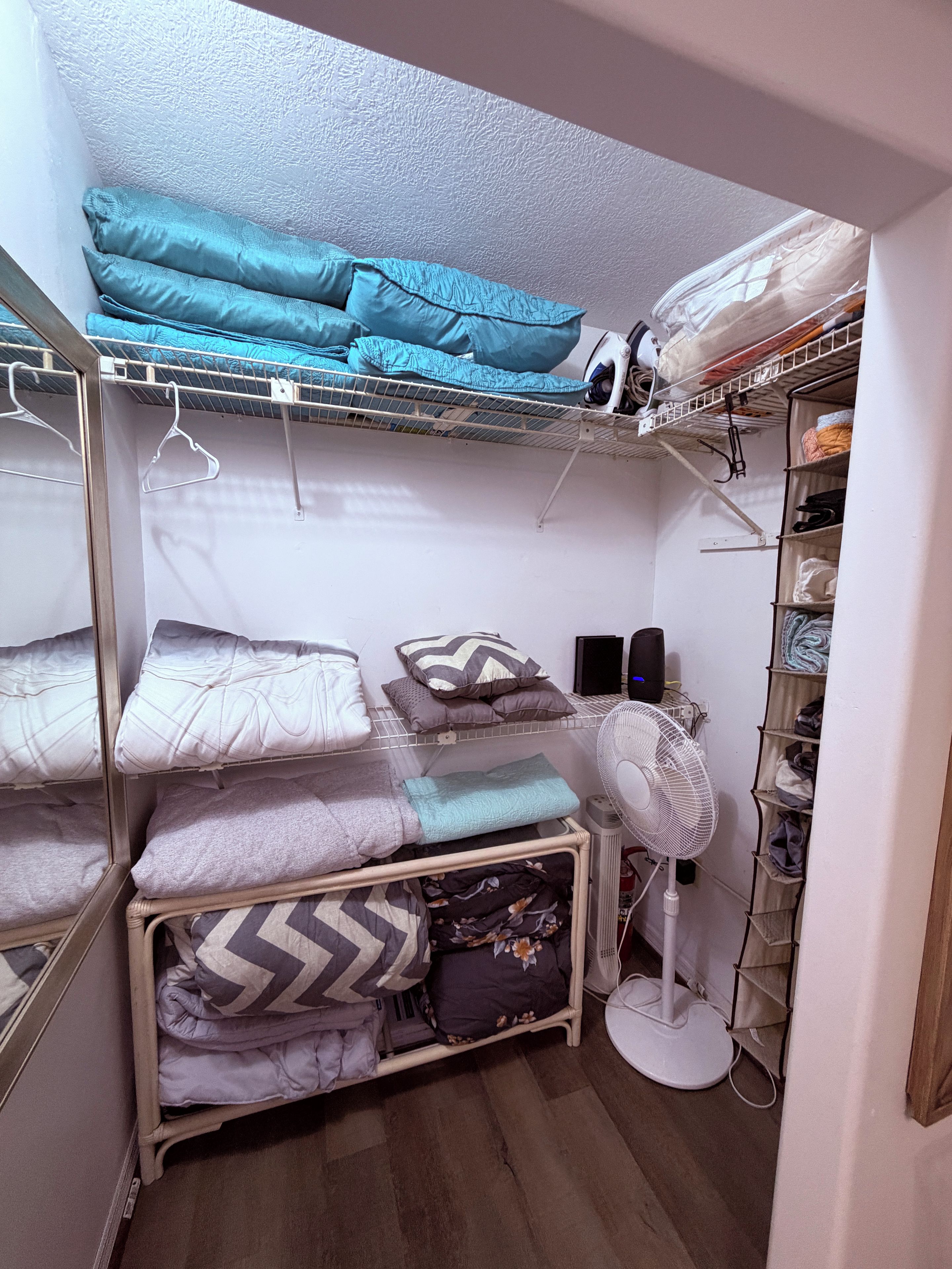 Un petit espace de rangement équipé d’étagères, d’un ventilateur et de linge de lit soigneusement plié.