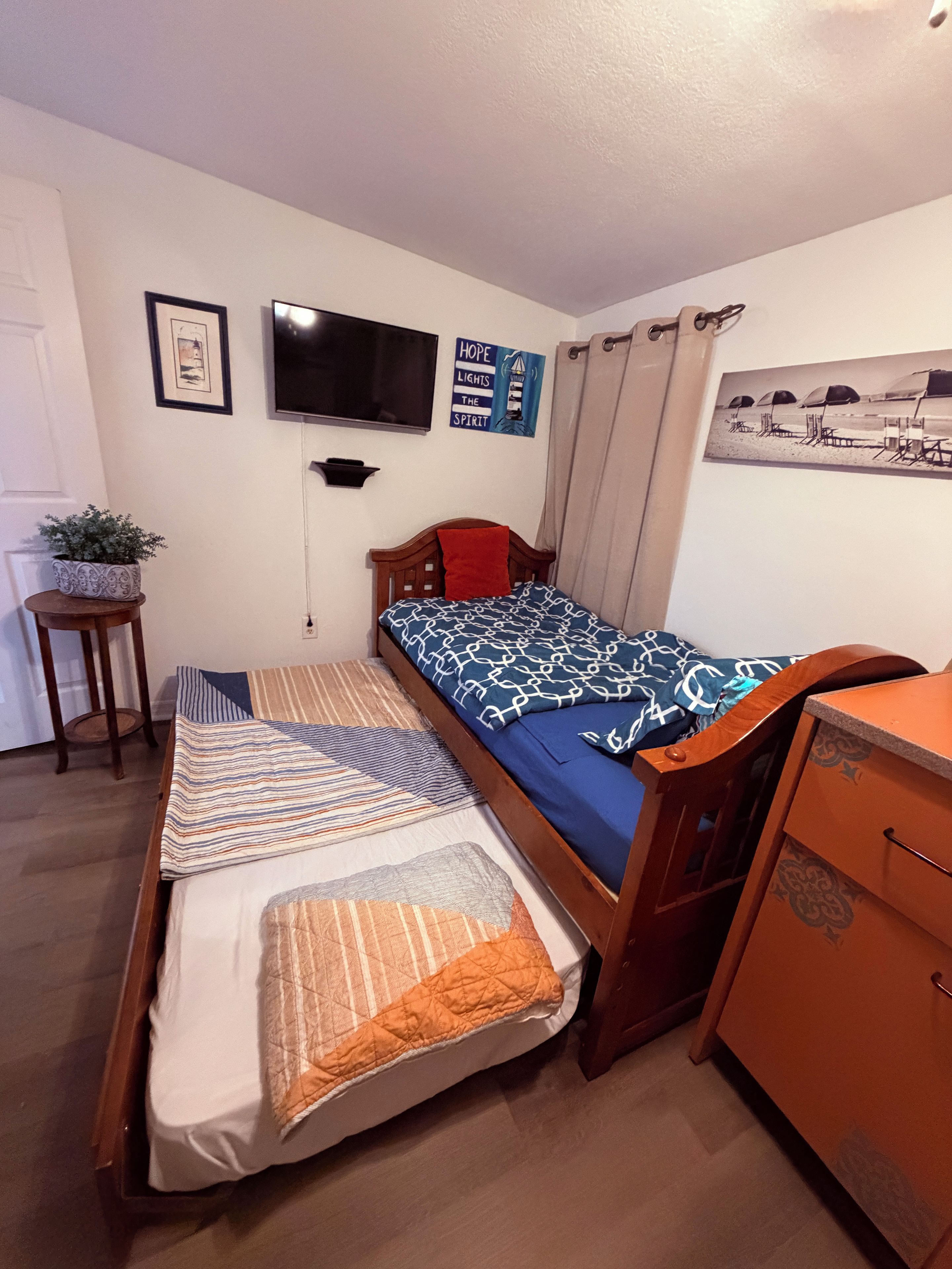 Een enkel bed met houten frame, een nachtkastje met lamp, een kleine bloempot, een tv aan de muur en een ingelijst foto.