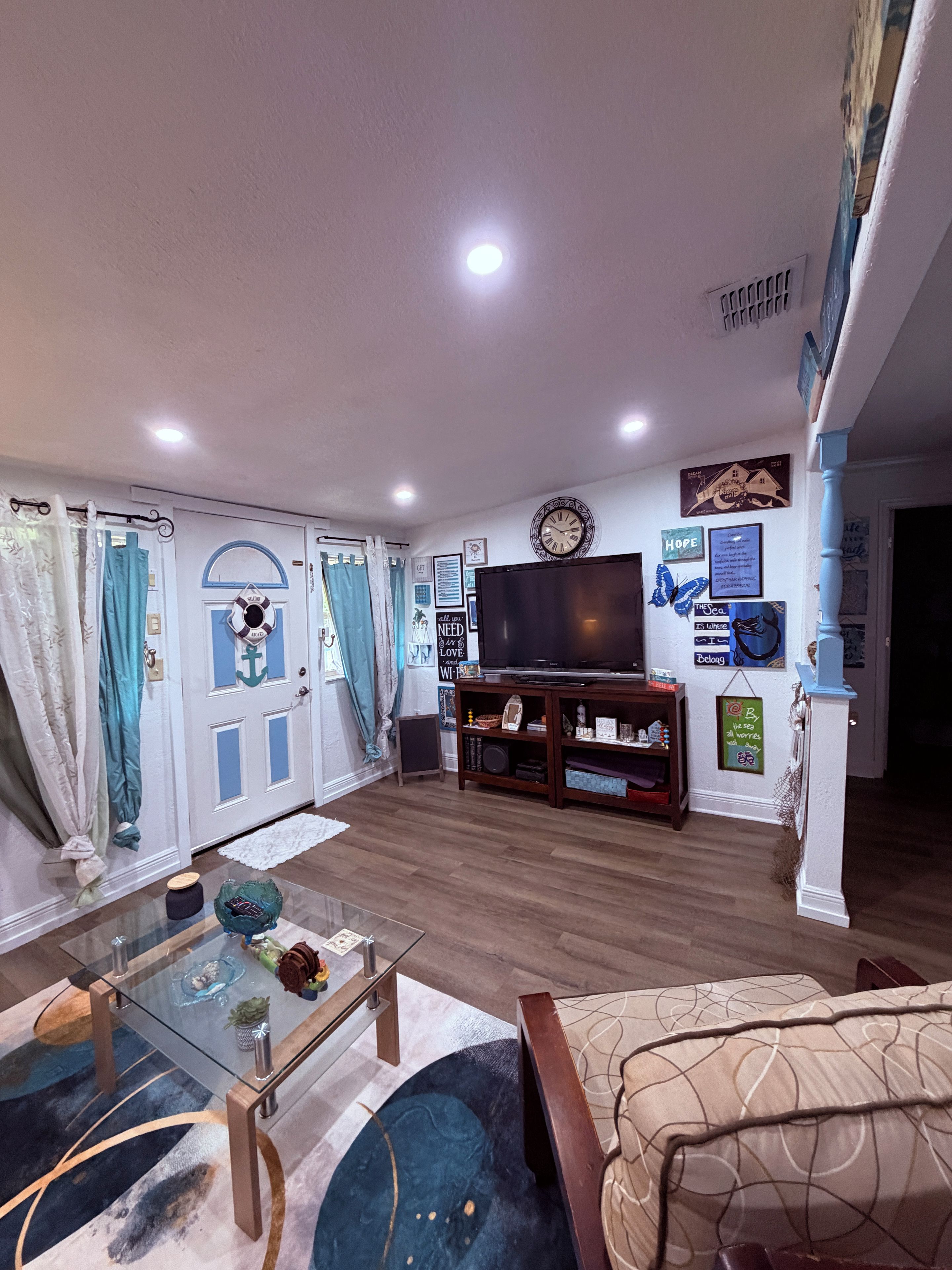 A living room with a flat-screen TV, wooden flooring, a glass coffee table, and a sofa.