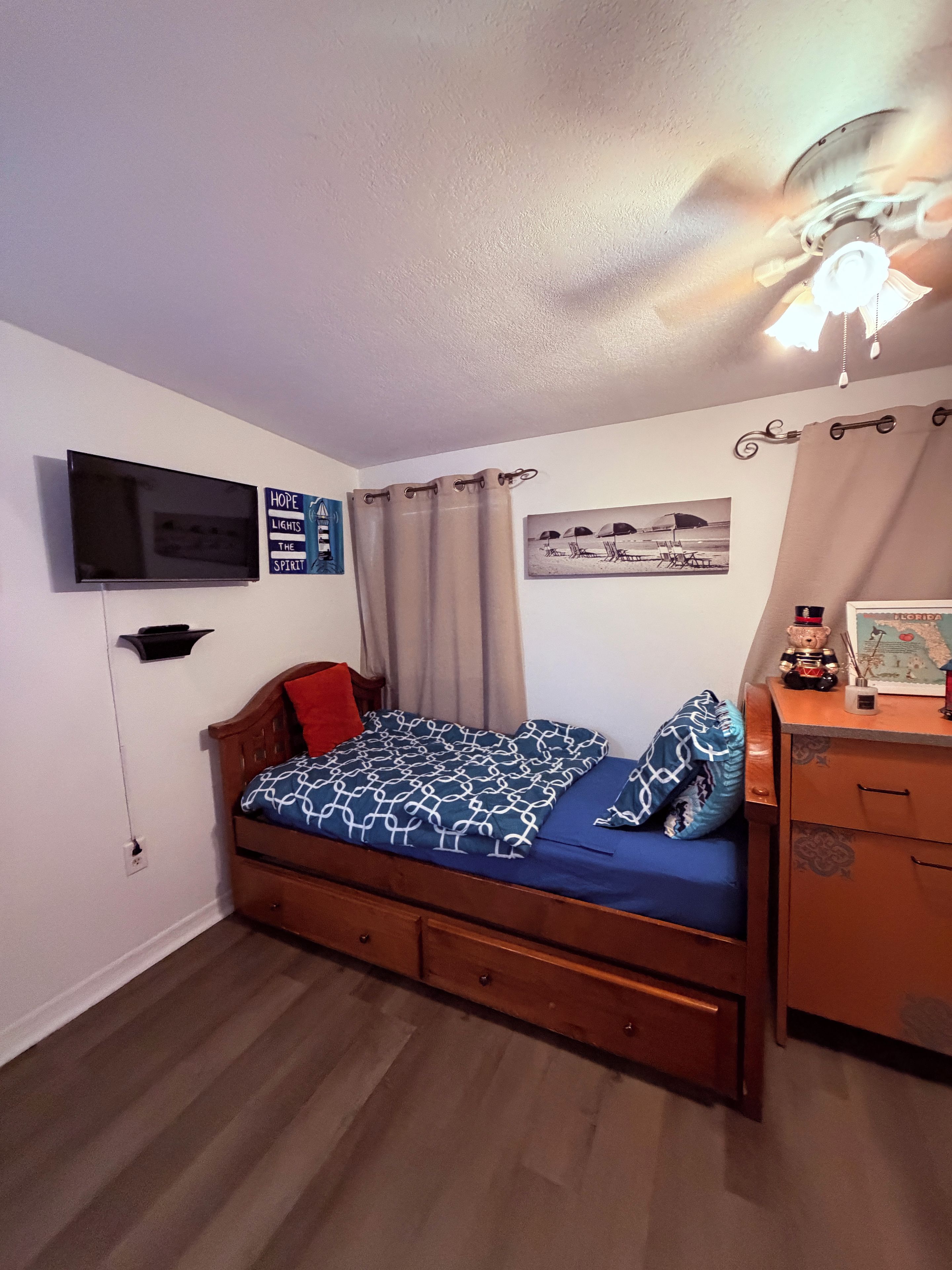 Une chambre à coucher comprenant un lit, un téléviseur, une étagère fixée au mur, un ventilateur de plafond et une carte posée sur la commode.