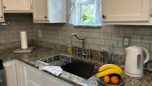 Uma cozinha com bancada de granito, pia com escorredor, janela com cortinas brancas e chaleira.
