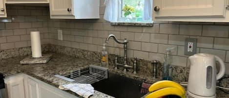 Uma cozinha com bancada de granito, pia com escorredor, janela com cortinas brancas e chaleira.