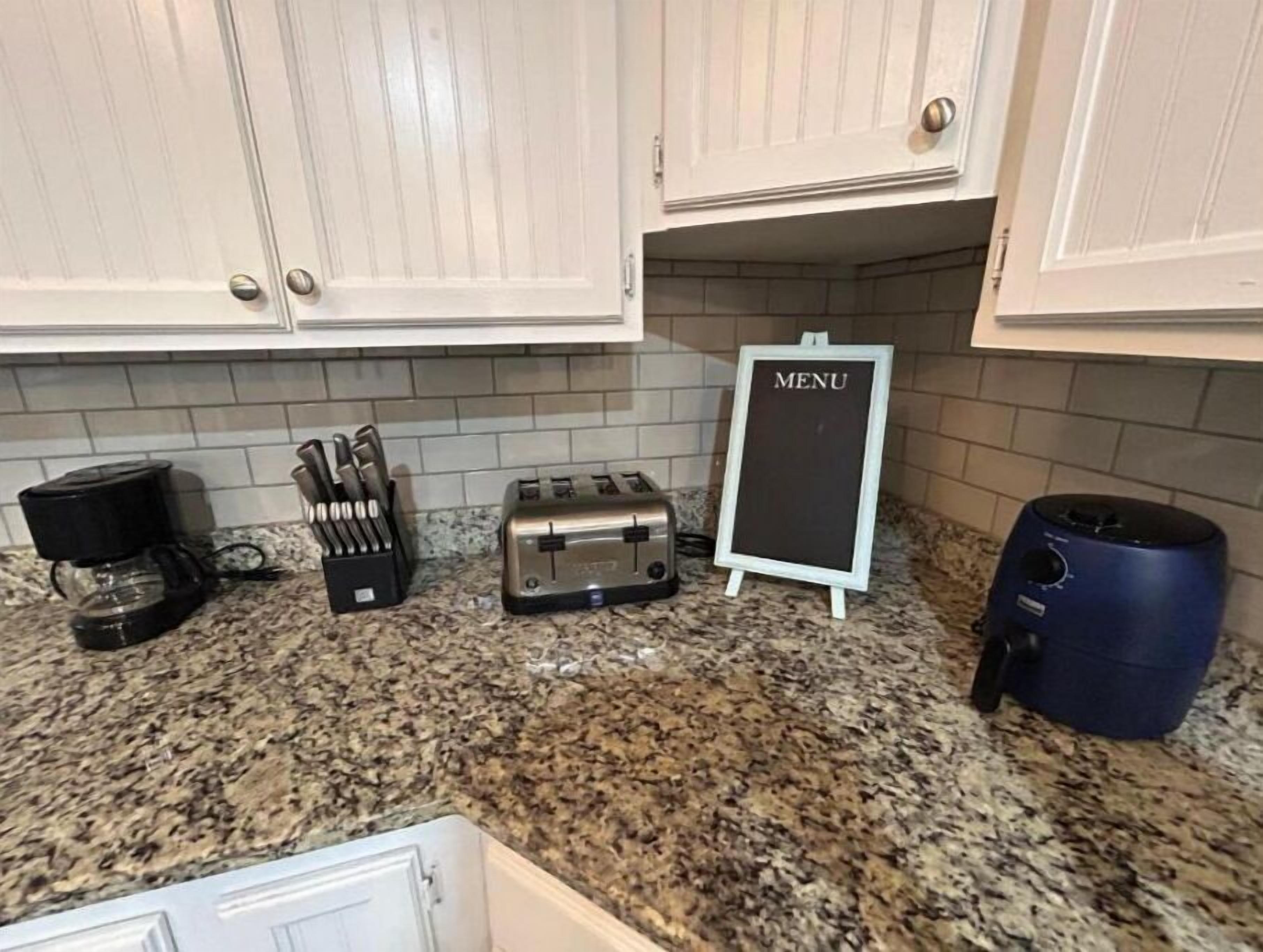 A kitchen with white cabinets, a granite countertop, a coffee maker, a toaster, a knife block, a menu board, and a blue appliance.