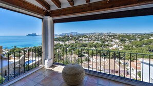 A balcony with a stone floor, a round stone bench, and a view of a coastal town with buildings and a mountain in the distance. - Bahía del Portet - By Almarina Villas (MORAIRA)