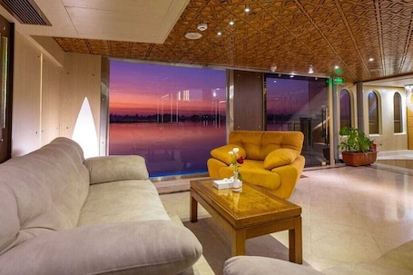 A modern lobby with a sofa, armchair, and coffee table. A large window offers a view of a sunset over a body of water.