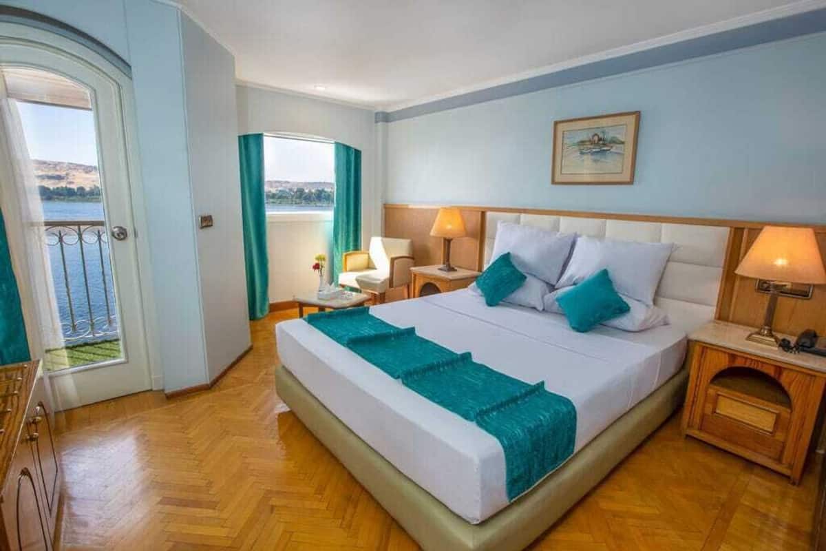 Una habitación de hotel con cama, mesitas de noche, vistas al exterior y una puerta que da a un balcón.