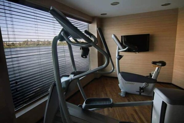 A gym with exercise equipment, a TV mounted on the wall, and a window with blinds.