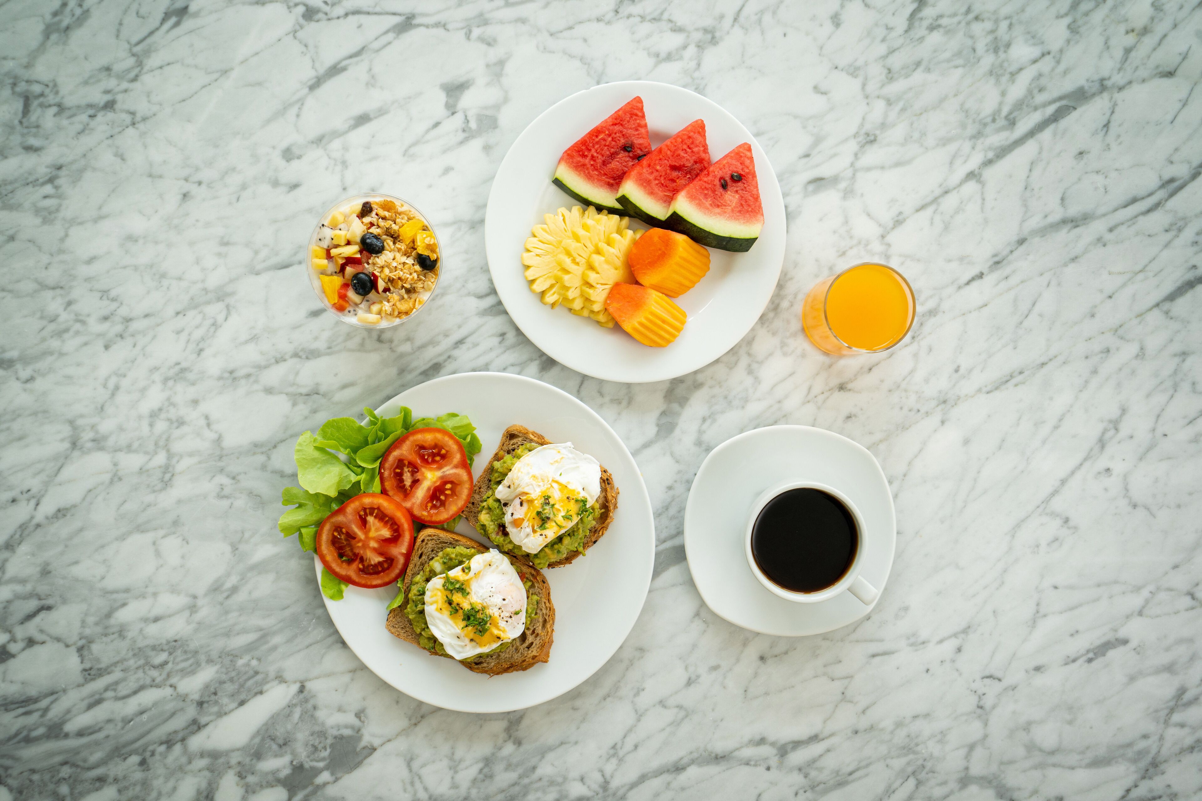 En morgenmadsanretning med ristet brød, æg, tomater, salat, vandmelon og et glas appelsinjuice på et marmorbord.
