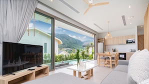 A modern living room with a flat-screen TV, a sofa, a dining table, and a kitchen area with wooden cabinets and a view of a mountain landscape. - Himmapana Villas - Hills  (Kamala)