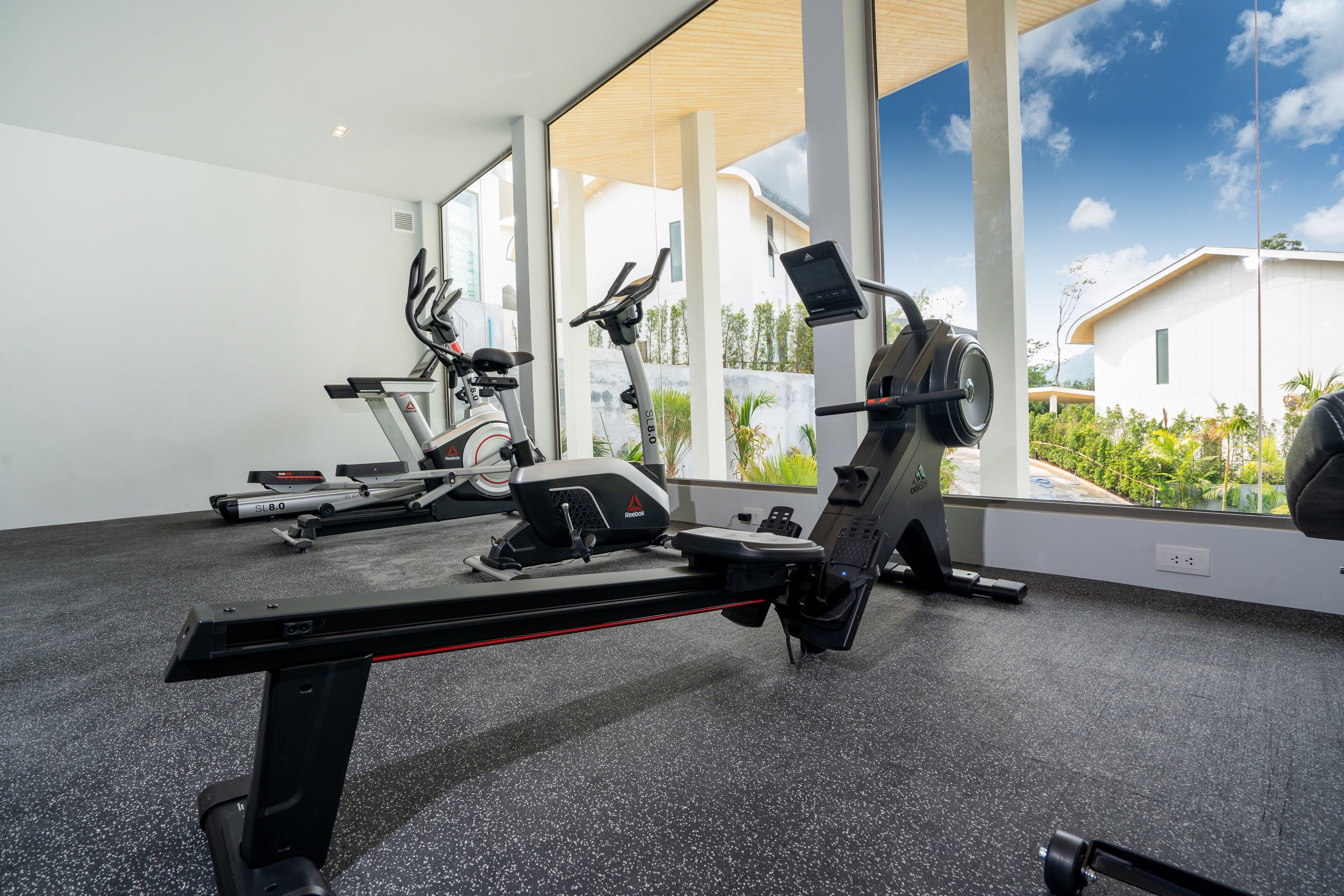 Un gimnasio con equipamiento de ejercicio, ventanas amplias y vistas a un edificio y zonas verdes.